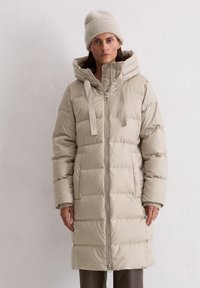 Nainen, joka pukeutunut vaaleanbeigeen vuorattuun pitkään takkiin, jossa on huppu, ja hänellä on päässään sopiva neulottu hattu, seisoo tasaista valkoista teksturoitua seinää vasten.