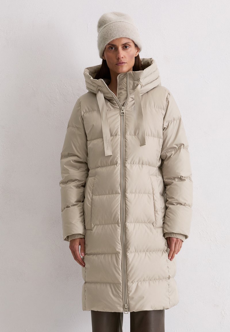 Nainen, joka pukeutunut vaaleanbeigeen vuorattuun pitkään takkiin, jossa on huppu, ja hänellä on päässään sopiva neulottu hattu, seisoo tasaista valkoista teksturoitua seinää vasten.