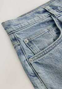 Jean en denim bleu clair avec coutures visibles, rivets métalliques et une petite poche avant sur un fond clair uni.