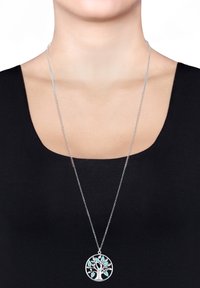 Collier en argent avec un pendentif en forme d'arbre, orné d'accents en turquoise, suspendu à une fine chaîne, porté sur un haut sombre.