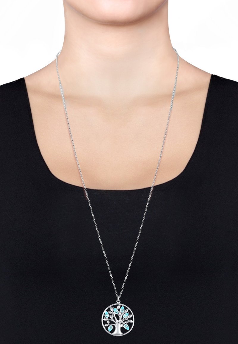Collier en argent avec un pendentif en forme d'arbre, orné d'accents en turquoise, suspendu à une fine chaîne, porté sur un haut sombre.