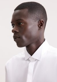 Camicia bianca con colletto a punta, apertura con bottoni, texture liscia e vestibilità aderente, mostrata di profilo su uno sfondo chiaro.
