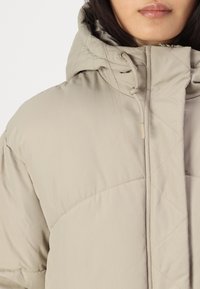 Casaco puffer bege claro com capuz, apresentando um padrão matelassado, cordão elástico e um acabamento liso e mate.