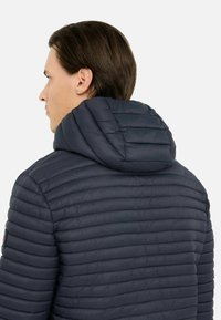 Tamsiai mėlynas pūkinis džemperis su horizontaliomis siūlėmis ir apvalia, paminkštinta kapuce; ant kairiojo rankovės yra guminis logotipo akcentas.