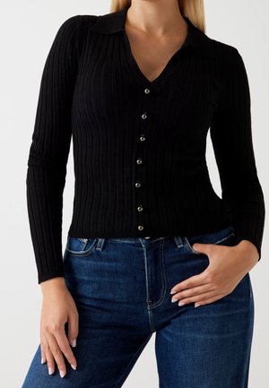 Femme portant un cardigan noir côtelé boutonné à manches longues et un jean bleu, une main dans la poche et l'autre détendue.