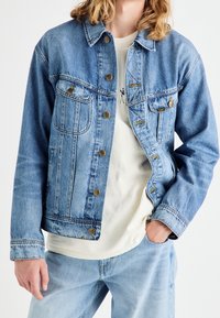 Ljusblå denimjacka med krage, framfickor och mässingsknappar. Bärs över en vit skjorta och matchas med ljusblå jeans.