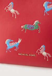 Κόκκινη δερμάτινη επιφάνεια με πολύχρωμες απεικονίσεις αλόγων και χρυσό λογότυπο "Michael Kors" στο κέντρο κοντά στο κάτω μέρος.