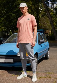 Vaaleanpunainen puuvillapaita, jossa on pieni logo, harmaat collegehousut sivuyksityiskohdilla ja valkoiset lenkkarit vihreillä yksityiskohdilla. Taustalla on sininen auto.