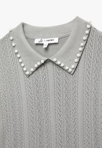 Suéter gris de punto con una textura estampada, un cuello con solapa y acentos decorativos de perlas a lo largo del collar. Etiqueta de la marca visible.