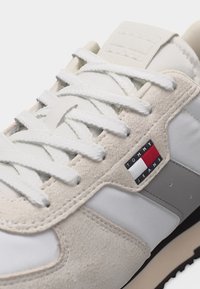 Gros plan sur une basket beige et blanche avec des lacets blancs et le logo Tommy Jeans sur le panneau latéral en daim.