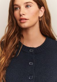 Cardigan a maglia blu navy con collo rotondo, caratterizzato da quattro bottoni scuri e un tessuto testurizzato. Include capelli lunghi e ondulati e orecchini a cerchio.
