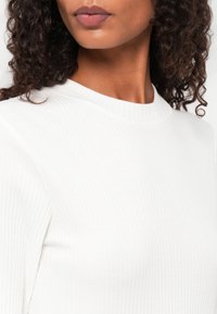 Hvitt ribbet topp med høy hals, som har en glatt tekstur og tettsittende design, delvis vist fra kragen og ned.