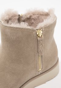 Botín de ante beige hasta el tobillo con cremallera de hardware dorado, cuello de borreguillo y suela lisa. Presenta costuras visibles y un suave forro interior.