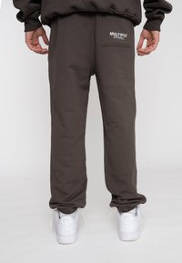 Person in braunen Jogginghosen mit dem Schriftzug "MULTIPLY APPAREL" auf der Gesäßtasche und weißen Nike Air Sneakers, stehend vor weißem Hintergrund.