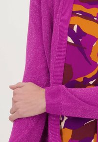 Fuchsia cardigan gemaakt van een sprankelend materiaal, met geribbelde accenten en een open voorkant ontwerp, gedragen over een kleurrijk patroon top eronder.