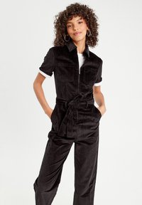 Black corduroy jumpsuit with short sleeves, front pockets, and a belted waist. Features a collared neckline and contrasting white cuffs.