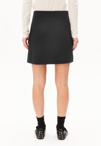 Frau, die mit dem Rücken steht, trägt einen schwarzen Minirock, ein weißes Langarmshirt, schwarze Socken und schwarze, mit Nieten besetzte Schuhe vor einem weißen Hintergrund.