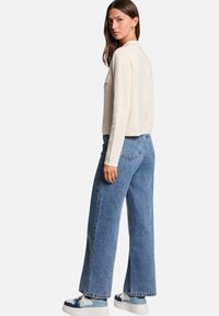 Haut tricot crème à manches longues associé à un jean en denim bleu à taille haute et jambes larges, ainsi qu'à des baskets bleu et blanc. Texture douce, coupe décontractée.