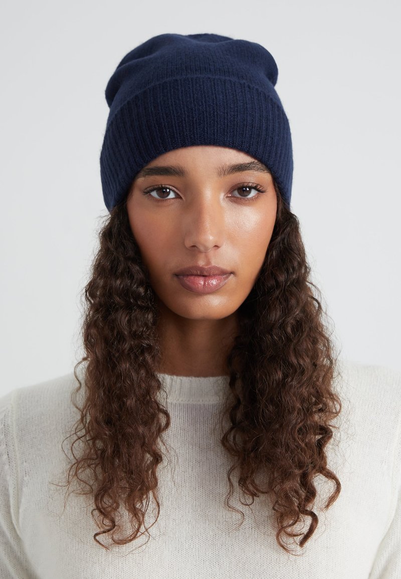 Bonnet tricoté marine avec une texture côtelée, un design légèrement décontracté et une finition lisse. Porté avec un pull de couleur claire.