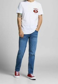 Muž na sobě má bílé tričko San Francisco 49ers, modré džíny, červené tenisky, s tetováním na levém rameni a pravou rukou v kapse, stojící na jednolitém pozadí.