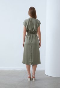 Olive green satin dress with short sleeves, cinched waist, and a knee-length design. Features a small back keyhole and smooth texture.