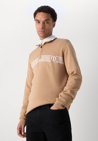 Beżowy sweter z dzianiny z zamkiem przy kołnierzu, z białymi poziomymi paskami i emblematem na klatce piersiowej. W zestawieniu z czarnymi spodniami.