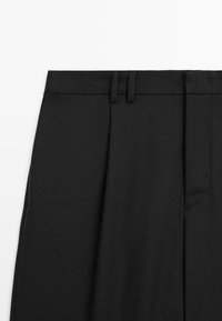 Pantalon noir ajusté avec un devant plat, des plis avant doubles et des poches latérales en tissu texturé.