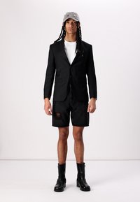 Sort skræddersyet blazer over en hvid t-shirt, parret med slidte sorte denimshorts og sorte ankelstøvler. Grå kasket med udsmykning.