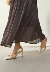 Zelta augstpapēžu kurpes ar norādītu purngalu un mazu loku detaļu, valkātas ar plūstošu tumšu caurspīdīgu svārku, kas izceļ sevī elegantu dizainu.