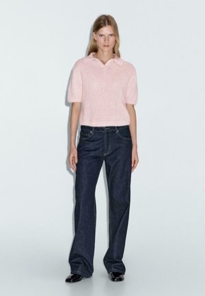 Maglione rosa chiaro a maniche corte con colletto, realizzato in tessuto morbido. Indossato con jeans a zampa d'elefante blu scuro e scarpe nere. Design semplice.
