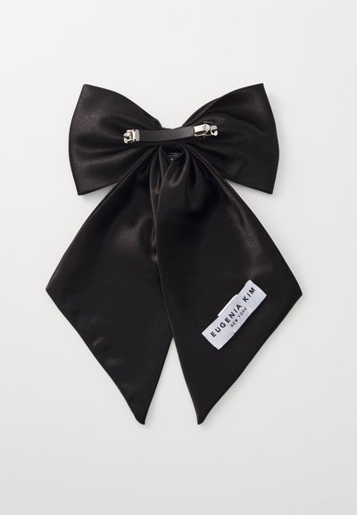 Svart satengsløyfe med to lange haler, festet med en sølvklips. Inkluderer en hvit etikett med "Eugenia Kim New York" merkevare.