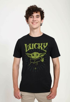 Sort bomulds T-shirt med et grønt grafisk motiv af en karakter med store ører og ordet "LUCKY" ovenfor, sat mod et burst-mønster.