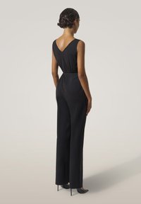 Kvinna med kort lockigt hår klädd i en ärmlös svart jumpsuit med V-rygg och svarta högklackade skor, stående med ryggen vänd framåt.