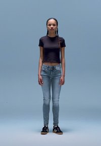 Svart cropped t-skjorte, lyseblå skinny jeans, svarte joggesko med hvite striper. Stående positur mot en ensfarget lyseblå bakgrunn.