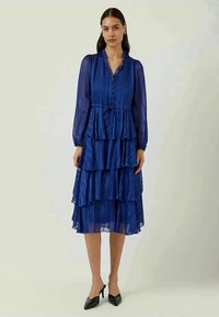 Μπλε, tiered midi φόρεμα με διάφανο ύφασμα, ριγέ λαιμόκοψη, κουμπιά, μακριά μανίκια και ζώνη με κορδόνι. Φοριέται με μαύρες γόβες.