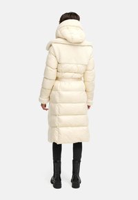 Creme pufferjakke med faux shearling krave, mellemlang længde, bælte i taljen og quiltet design. Model iført sorte leggings og støvler.