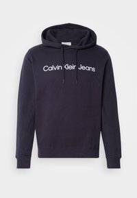 Mörkblå huvtröja i bomullsblandning med en framficka i kängurustil. Har en vit "Calvin Klein Jeans"-logotyp över bröstet och dragsko i huven.