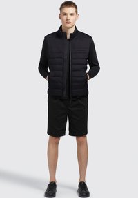 Sort puffervest med lynlås foran, parret med en sort T-shirt med korte ærmer og sorte shorts. Iført med sorte sneakers.