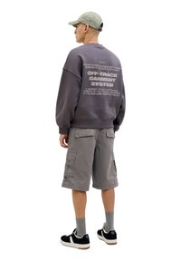 Gray sweatshirt with back text, paired with gray cargo shorts. Model wears black sneakers with white accents and gray socks, standing in profile.