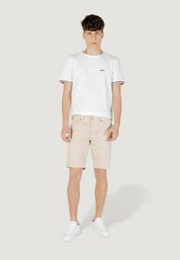 T-shirt bianca in cotone con logo nero, bordo a contrasto nero. Pantaloni beige, con tasche e chiusura a bottone. Sneakers bianche con suole piatte.