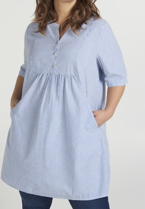 Femme portant une tunique ample à rayures verticales bleues et blanches avec manches mi-longues, boutons et mains dans les poches.