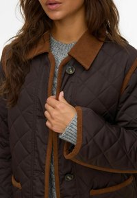 Giacca trapuntata marrone scuro con colletto e dettagli in velluto, dotata di chiusura a bottoni e tasche laterali. Maglione grigio visibile sotto.
