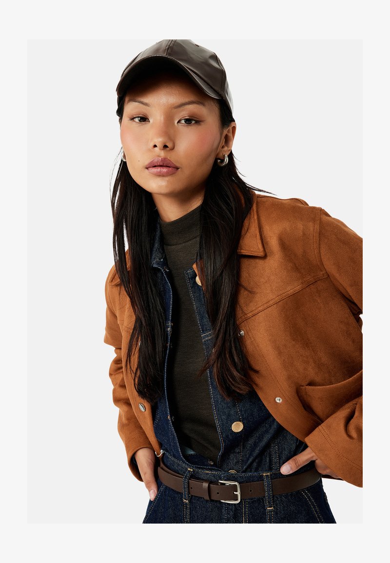 Ung kvinde med langt mørkt hår iført en brun læderkasket, brun ruskindsjakke, mørk rullekrave og denim-dragter med brun bælte.