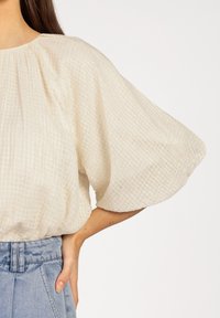 Romige blouse met een brede, vloeiende mouw en een geplooide halslijn, gecombineerd met lichtblauwe, high-waisted jeans.