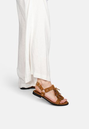 Pied portant une sandale en daim marron à franges avec des pampilles perlées, bride à la cheville et boucle dorée, associée à un pantalon blanc ample.