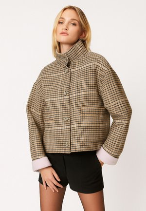 Donna che indossa una giacca beige a quadri con bottoni, collo alto e maniche arrotolate, abbinata a pantaloncini neri e calze trasparenti.