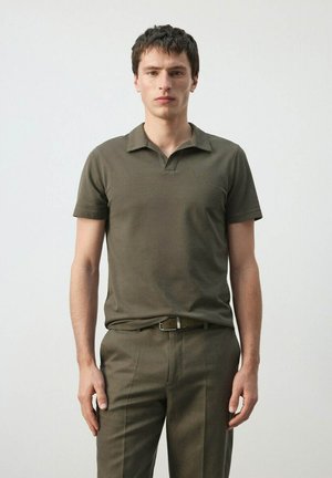 Joven de pie con camiseta tipo polo verde oliva y pantalones a juego con cinturón, frente a un fondo claro y liso.