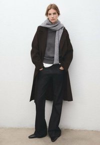 Loszittende donkerbruine jas, grijze gebreide trui, wit overhemd, op maat gemaakte donkere broek, grijze sjaal, gestroomlijnde zwarte schoenen, gestructureerde oppervlakken.