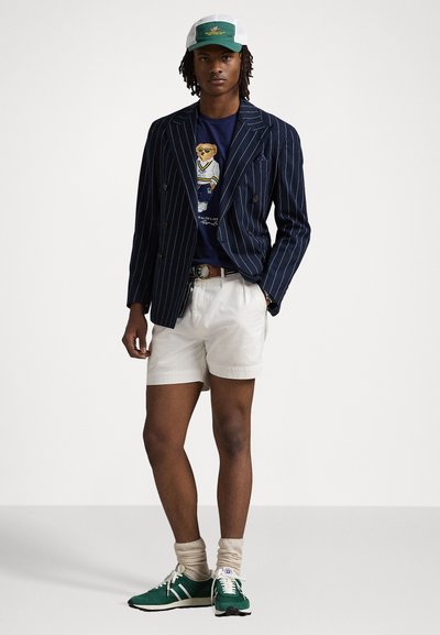Marine gestreepte blazer over een graphic t-shirt, witte shorts, groene sneakers en een groen/witte pet. Heeft een relaxte pasvorm en contrasterende kleuren.