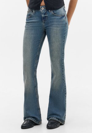 Nő kék csíkos bootcut farmert visel fekete cipővel, fehér háttér előtt áll.